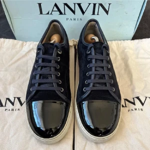 Lanvin svarta sneakers med lackad tå - Snygga svarta sneakers från Lanvin med ovandel i mocka och en glansig, lackad tå. Skorna har vita platta sulor och svarta snören. Klassisk och stilren design som sticker ut med den blanka tån. Perfekta för dig som vill ha något unikt och exklusivt.