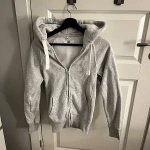 Russemerch hoodie med dragkedja XS - Ljusgrå hoodie i storlek XS från R med dragkedja framtill, huva med snörning och två fickor. Mjuk insida och ribbade muddar vid ärmslut och nederkant. Perfekt för chill dagar eller när du vill vara extra bekväm.