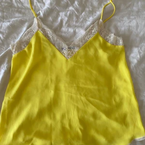 Gul linnetopp med spetsdetalj - Snygg gul linnetopp med tunna axelband och vit spets längs v-ringningen. Toppen har en slät, glansig yta och är perfekt för varma dagar. Den har en feminin och fräsch look med fina detaljer upptill.