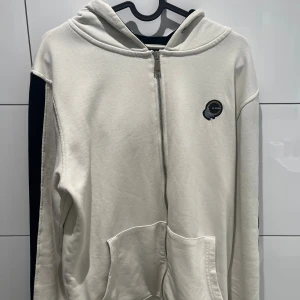 Vit hoodie med dragkedja från La Vichy - Snygg vit hoodie från La Vichy i storlek M med hel dragkedja framtill, huva och broderad logga på bröstet. Tröjan har fickor framtill och svarta detaljer längs ärmarna. Perfekt för en chill och stilren look.