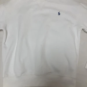 Vit sweatshirt från Polo Ralph Lauren - Vit sweatshirt från Polo Ralph Lauren med klassisk blå broderad logga på bröstet. Tröjan har rund halsringning, ribbade muddar och är långärmad. Perfekt för en clean och stilren look.