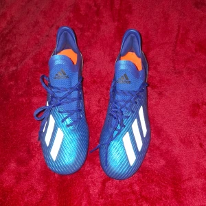 blåa Adidas Nemesis fotbollsskor  - Säljer ett par blåa Adidas Nemesis fotbollsskor med vita ränder och svarta detaljer vid hälen. Skorna har snörning, platt sula med dobbar och orange innersula. Tillverkade i syntetmaterial och designade för att ge bra grepp på planen.