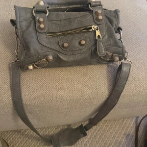Grå axelväska med detaljer - Säljer en grå axelväska i skinn med coola nitar och dragkedja framtill. Väskan har både handtag och lång axelrem, samt flätad detalj på handtaget. Perfekt storlek för det viktigaste och har flera fack inuti.