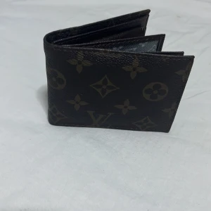 Louis Vuitton plånbok i skinn - Snygg plånbok från Louis Vuitton i mörkbrunt skinn med klassiskt monogrammönster. Kompakt och smidig modell med plats för kort och sedlar. Perfekt accessoar för dig som gillar lyxiga detaljer och tidlös design.(nytt kostar runt 5000kr