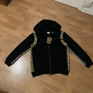Exklusiv Burberry Hoodie - svart med rutmönster - Svår att få tag på! En exklusiv Burberry hoodie i svart med dragkedja, prydd med märkets klassiska rutmönster längs ärmar och sidor. Bekväm passform, ett måste i garderoben för dig som vill sticka ut med stil. Perfekt mix av lyx och streetwear, säljs inte ofta!  Storlek - L  Skick - Nytt skick, oanvänd