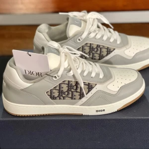 Dior B27 sneakers i grått och vitt - Snygga Dior B27 sneakers i vitt och ljusgrått skinn med klassiskt Dior-mönster på sidorna. Skorna har vita snören, perforerad tå och gummisula med logga på hälen och sidan. Perfekt för dig som vill ha en exklusiv och trendig look.