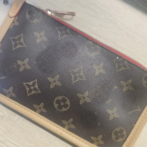 monogram kuvertväska - Snygg kuvertväska med klassiskt monogrammönster i brunt och beige. Väskan har guldfärgad dragkedja och röda detaljer längs kanten. Perfekt för att bära det viktigaste med stil.