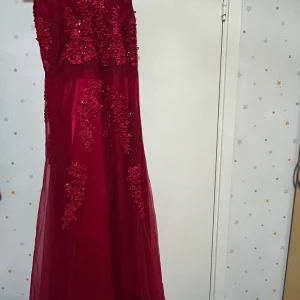 Vinröd långklänning med spetsdetaljer - Säljer en vinröd långklänning med v-ringning och vackra spetsapplikationer samt paljetter upptill. Klänningen har en flowy kjol i tunt tyg och markerad midja. Perfekt för bal eller festliga tillfällen när du vill sticka ut.