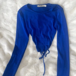 Blå croppad topp med snörning NA-KD - Säljer en klarblå croppad topp från NA-KD med långa ärmar och snygg snörningsdetalj i ryggen. Toppen har en tight passform och är tillverkad i ett mjukt, stretchigt material som sitter skönt på kroppen. Perfekt för att lyfta din outfit med en pop av färg.