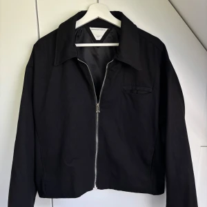 Svart jacka från Bottega Veneta - Svart jacka från Bottega Veneta med klassisk krage och silverfärgad dragkedja framtill. Jackan har en minimalistisk design med en liten bröstficka och är tillverkad i ett slätt tyg med fodrad insida. Perfekt för dig som gillar stilren och exklusiv look. INTE ÄKTA