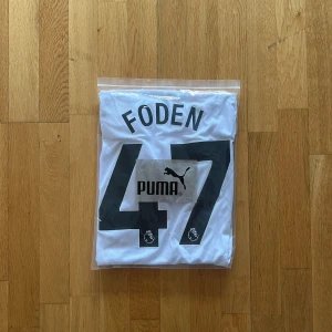 Manchester City Foden Puma fotbollströja - Snygg Manchester City fotbollströja från Puma med Foden och nummer 47 på ryggen. Tröjan är vit med grått mönster, har en diagonal svart och röd rand på framsidan samt klubbmärke och sponsortryck. Tillverkad i lätt RE:FIBRE-material, perfekt för match eller träning.