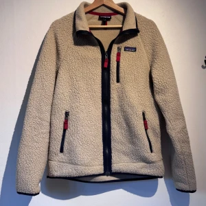 Beige teddyjacka från Patagonia - Mysig beige teddyjacka från Patagonia med svart dragkedja och röda detaljer på fickorna. Jackan har hög krage, tre fickor med dragkedja och långärmad design. Perfekt för kyliga dagar och har en skön, fluffig känsla.