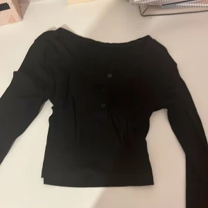 Svart långärmad topp från Dazy - Svart långärmad topp från Dazy med rund hals och tre dekorativa knappar framtill. Modellen är ribbad och har en slim passform som sitter snyggt på kroppen. Perfekt att styla med jeans eller kjol för en clean look.