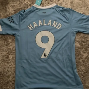 Manchester City Haaland tröja Puma S - Manchester Citys officiella matchtröja från Puma med Haaland och nummer 9 på ryggen. Tröjan är ljusblå med vita detaljer, klubbmärke på bröstet och diagonal rand. Tillverkad i lätt och ventilerande polyester, perfekt för fotboll eller supporterliv.