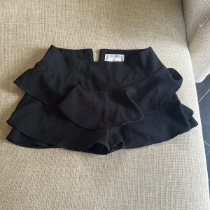 Svarta volangshorts med dragkedja - Säljer ett par svarta shorts med dubbla volanglager och hög midja. De har en dragkedja bak för enkel påtagning och är tillverkade i ett mjukt, vävt tyg. Perfekta för dig som gillar en trendig och lekfull look. Passar om du har storlek S i underdel