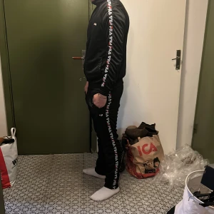 Svart FILA ziptröja och byxor med logga - Svart ziptröja och byxor från FILA med logga på bröstet och längs ärmarna och benen. Tröjan har lång ärm, ribbade muddar och dragkedja framtill. Tillverkad i glansig polyester, perfekt för träning eller chill. Snygg och sportig look med klassisk FILA-design.