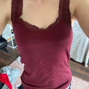 Vinröd linnetopp med spets från H&M - Säljer en vinröd linnetopp från H&M med breda axelband och fina spetsdetaljer längs ringningen och banden. Toppen är ärmlös och har en mjuk, stretchig känsla som gör den superbekväm. 