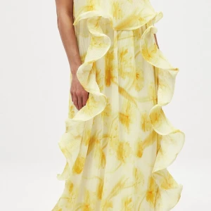 Gul blommig långklänning med volang - Långklänning i ljusgul nyans med blommigt mönster i gult. Klänningen har breda volangdetaljer längs hela framsidan. Den är aldrig använd.