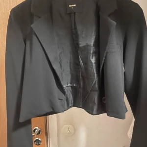 Svart croppad kavaj från Bik Bok - Snygg svart croppad kavaj från Bik Bok med klassisk krage och knapp framtill. Jackan har lång ärm och är fodrad på insidan för extra komfort. Perfekt för dig som vill ha en trendig och stilren look.