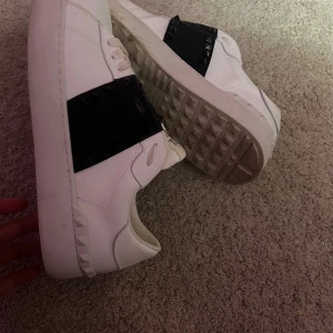 Valentino Garavani Rockstud sneakers - Snygga vita Valentino Garavani Rockstud sneakers i skinn med svart band och ikoniska nitar över vristen. Klassisk rund tå, platt sula och vita snören. Perfekta för dig som vill ha en lyxig och edgy touch till din outfit.