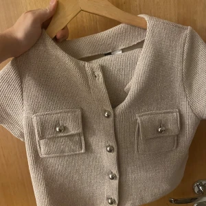 Beige stickad croppad  med fickor och tillhörande kjol - Säljer en beige stickad med korta ärmar och croppad passform med tillhörande kjol. Toppen har två snygga fickor framtill med silvriga knappar och en rad knappar längs hela framsidan. Perfekt för dig som gillar trendiga detaljer och neutral färg.