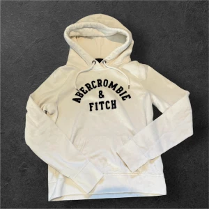 Vit hoodie från Abercrombie & Fitch - Vit hoodie från Abercrombie & Fitch med svart broderad logga på bröstet. Tröjan har huva med snörning, känguruficka och långa ärmar. Mjuk insida och klassisk passform, perfekt för en avslappnad stil.