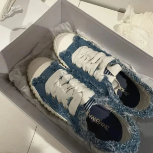Zimmermann denim sneakers med fransar - Unika sneakers från Zimmermann i ljusblå denim med fransade kanter och breda vita snören. Skorna har en chunky, räfflad sula i beige och coola detaljer som ger en avslappnad streetstyle-vibe. Perfekta för dig som gillar statement-skor och denim. Box medföljer och har även bild på kvitto och orderbekräftelse.