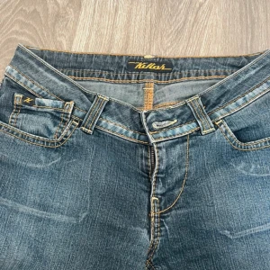 Blå lågmidjade bootcut jeans - Snygga blå jeans från Kilash med klassisk bootcut-modell. Jeansen har kontrastsömmar i gult, fem fickor och normal passform. Tillverkade i jeansmaterial med en lätt tvättad look och detaljerade bakfickor med broderad logga.
