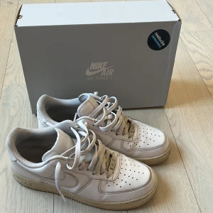 Nike Air Force 1 vita sneakers - Klassiska Nike Air Force 1 sneakers i helvitt skinn med perforerad tå, rund tå och platt sula. Skorna har snörning och ikonisk Nike Swoosh på sidorna. Perfekta för dig som gillar en clean och tidlös streetstil. Skick 7/10.
