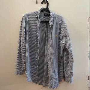Randig skjorta från Ralph Lauren - Säljer en klassisk blåvit randig skjorta från Ralph Lauren med gul broderad logga på bröstet. Skjortan har lång ärm, knappar framtill och är i bomull. Classic fit-modell som ger en snygg och tidlös look.