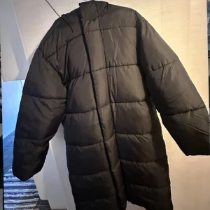 Svart lång pufferjacka med huva från asos - Säljer en svart pufferjacka med huva och långa ärmar. Jackan har ett quiltat mönster med breda rutor och stängs med både dragkedja och knappar. Perfekt för kalla dagar och har en oversized passform som ger en cool vibe.