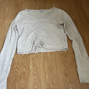 Beige ribbad croppad långärmad topp - Säljer en beige croppad topp med långa ärmar och snygg v-ringning. Toppen har ribbad struktur och en knytning framtill som ger en cool detalj. Perfekt för att styla med jeans eller kjol för en chill look.