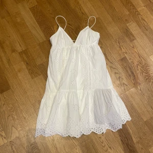 Vit somrig klänning med broderi - Säljer en slutsåld klänning från Zara med tunna axelband och v-ringad front. Klänningen har broderade blommor och spetsdetaljer, samt en volangkant nertill med hålmönster. Säljer då den tyvärr inte passar mig längre. Skriv för mer bilder eller frågor💕