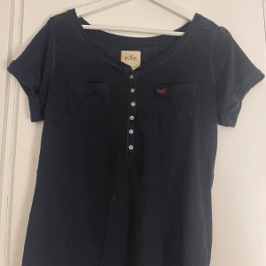 Svart t-shirt från Hollister med knappar - Svart t-shirt från Hollister med korta ärmar och rund hals. Framtill finns en knappslå med sex knappar och två bröstfickor, varav en har Hollisters röda logotyp.