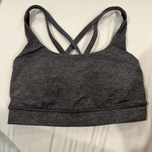 Mörkgrå sporttopp från Lululemon - Säljer en mörkgrå sporttopp från Lululemon med snygga korsade axelband i ryggen. Toppen är i stretchigt material och har en enkel, stilren design utan mönster. Perfekt för träning och aktiva dagar. Liten Lululemon-logga på ryggen. Storlek 2, använd ett par gånger bara.