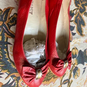 Röda ballerinaskor från Bally med rosett - Snygga röda ballerinaskor från Bally i skinn med rund tå och dekorativ rosett framtill med gulddetalj. Skorna har en låg kilklack i röd- och vitmönstrad textil som ger en unik touch. Perfekta för dig som vill sticka ut med klassisk stil.