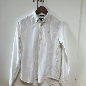 Vit slim fit skjorta från Ralph Lauren - Vit skjorta från Ralph Lauren i slim fit-modell, tillverkad i 100% bomull. Klassisk button-down krage och broderad logga på bröstet. Långärmad med knappar framtill och vid ärmslut. Perfekt för dig som gillar stilrena och tidlösa plagg.