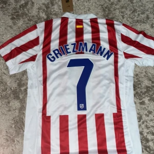 Atletico Madrid Griezmann tröja Nike S - Atletico Madrid matchtröja med Griezmann 7 på ryggen. Klassisk röd- och vitrandig design, officiell Nike Dri-Fit med klubbmärke och sponsortryck på bröstet. Skön och lätt polyester, perfekt för fotboll eller supporterliv.