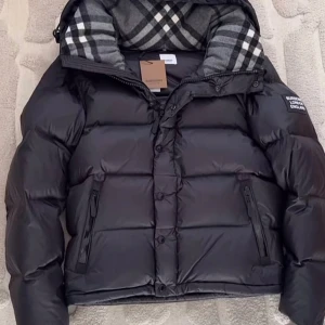 Svart pufferjacka från Burberry - Svart pufferjacka från Burberry med avtagbara ärmar så den kan användas som väst. Jackan har dragkedja och knappar framtill, två fickor med dragkedja och en huva med rutigt foder i grått och vitt. Stor Burberry-patch på ärmen.