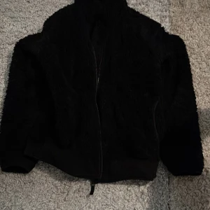 Svart teddyjacka från Uniqlo M - Mjuk och fluffig svart teddyjacka från Uniqlo i storlek M. Jackan har hel dragkedja framtill, hög krage och två sidofickor. Perfekt för dig som gillar en chill och cozy stil. Materialet är syntet och jackan har en loose passform. Den är även limited edition