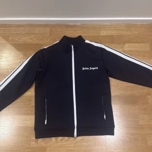 Svart track jacket från Palm Angels - Palm Angels svart track jacket med vita ränder längs ärmarna och vit dragkedja. Jackan har ståkrage, två fickor med dragkedja och Palm Angels-logga på bröstet. Materialet är mjukt och stretchigt, perfekt för en sportig streetwear-look.