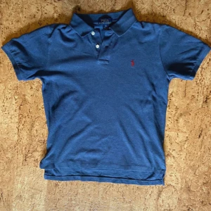 Blå pikétröja från Ralph Lauren - Klassisk blå pikétröja från Ralph Lauren med korta ärmar, krage och knappar framtill. Tröjan har det ikoniska röda broderade polospelar-loggan på bröstet och är gjord i mjuk bomull. Perfekt för en clean och stilren look.