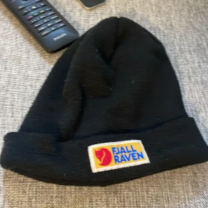 Svart mössa från Fjällräven - Svart mössa från Fjällräven med klassisk uppvikt kant och snyggt broderat märke framtill. Materialet är stickat och mjukt, perfekt för kyliga dagar. På sidan finns en liten svensk flagga som extra detalj. Enkel och stilren design som funkar till allt.
