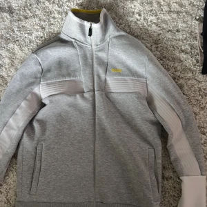 Grå kofta BOSS - Snygg grå vindjacka från BOSS med vita detaljer längs ärmarna och bröstet. Jackan har hel dragkedja framtill, hög krage och diskret gul BOSS-logga på bröstet. Perfekt för dig som gillar sportig och clean stil.