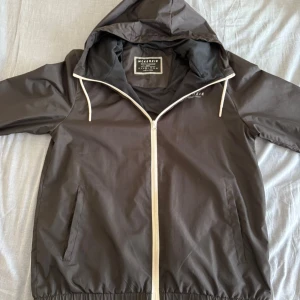 Svart vindjacka från McKenzie med huva - Svart vindjacka från McKenzie med vit dragkedja och snören i huvan. Jackan har två fickor framtill, elastisk nederkant och är helsvart med diskret logga på bröstet. Perfekt för blåsiga dagar och enkel att matcha med det mesta.