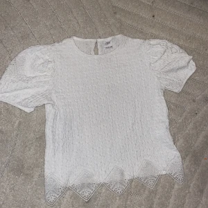 Vit blus med spetsdetaljer från Zara - Supersöt vit blus från Zara med puffiga korta ärmar och unik vågig struktur. Nederkanten har spetsdetaljer i geometriskt mönster som ger extra stilpoäng. Perfekt för dig som gillar romantiska och trendiga plagg med fina detaljer.