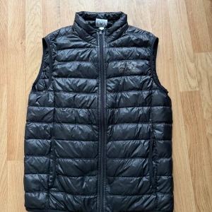Svart Emporio Armani EA7 - En svart stilig Emporio Armani EA7 väst med guldig liogga på vänstra bröstet med bra och äkta material. Skancod för att visa att den är äkta.