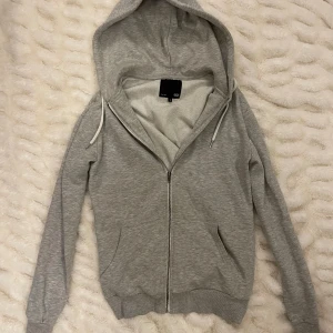 Grå zip hoodie - Grå  hoodie  med hel dragkedja framtill och huva med vita snören. Tröjan har två fickor på sidorna och ribbade muddar vid ärmslut och nederkant. Perfekt för chill dagar eller när du vill vara extra bekväm.