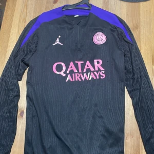 PSG långärmad fotbollströja Jordan - Snygg svart PSG fotbollströja från Jordan med lila axelpartier och lång ärm. Tröjan har Paris Saint-Germain-logga på bröstet, Jordan-logga och Qatar Airways-tryck i rosa och vitt. Tillverkad i lätt, ventilerande material med diskret mönster på ärmarna.