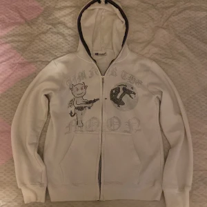 Vit hoodie Aim For The Moon XS - Vit hoodie med dragkedja från Aim For The Moon i storlek XS. Framsidan har ett tryck med en tecknad figur och texten 'Aim For The Moon' i silvriga detaljer. Tröjan har huva, långa ärmar och ribbade muddar. Perfekt för dig som gillar coola prints och streetstyle.